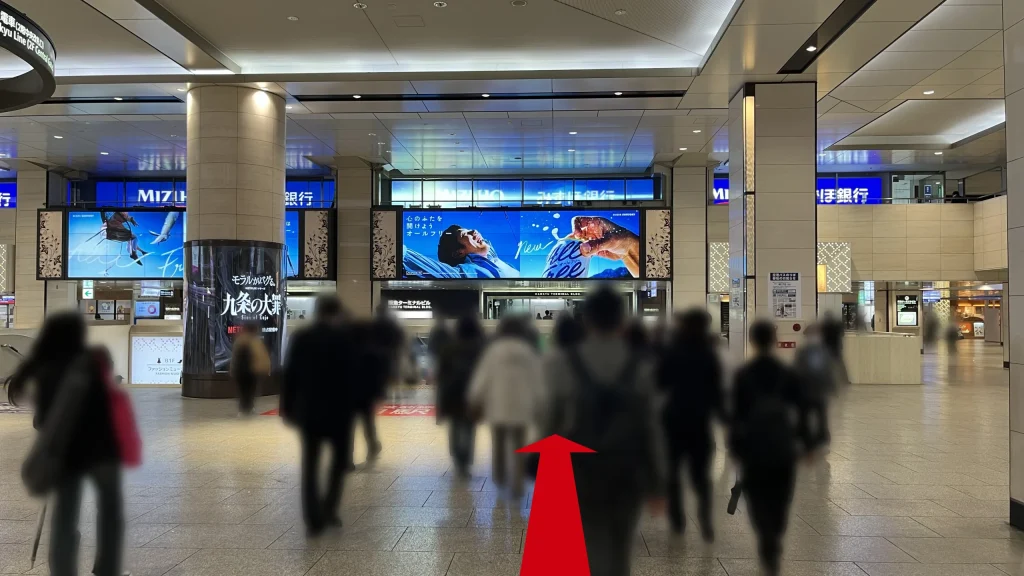 Alt:Signatureグラングリーンゲートタワーへのアクセス方法:エスカレーターを下りた先の1Fコンコースで、正面に「みずほ銀行」の看板や大型のデジタルサイネージがあり、多くの人々が歩いている様子。