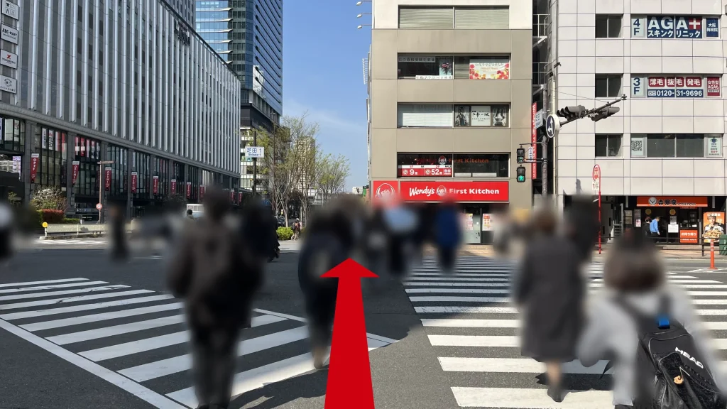 JAM BASEへのアクセス方法：「芝田1」の交差点の横断歩道の先にウェンディーズ・ファーストキッチンがあり、多くの歩行者が横断歩道を渡っている様子。
