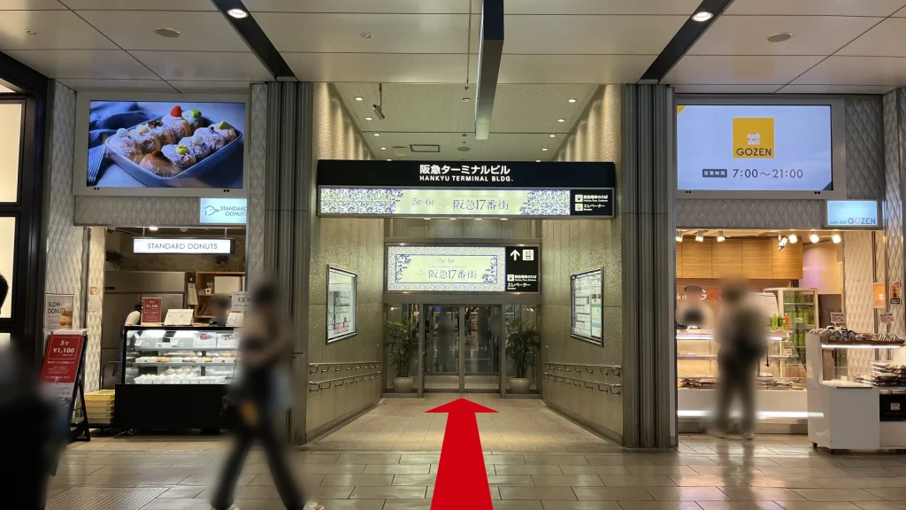 リージャス阪急ターミナルビルビジネスセンターへのアクセス方法:中央に「阪急ターミナルビル」と書かれた黒いサインボードがあり、その下に「5F-6F 阪急17番街」への案内があります。両サイドにはドーナツ店と惣菜店があります。