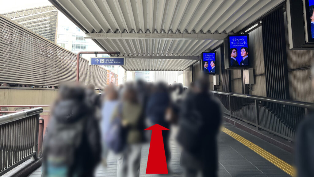 JAM BASEへのアクセス：「天ぷらまきの」の前を道なりに進んで屋外へ出た通路の図　前方に「↑JR大阪駅」と書かれた看板が見える　右手にはデジタルサイネージが並んでいる
