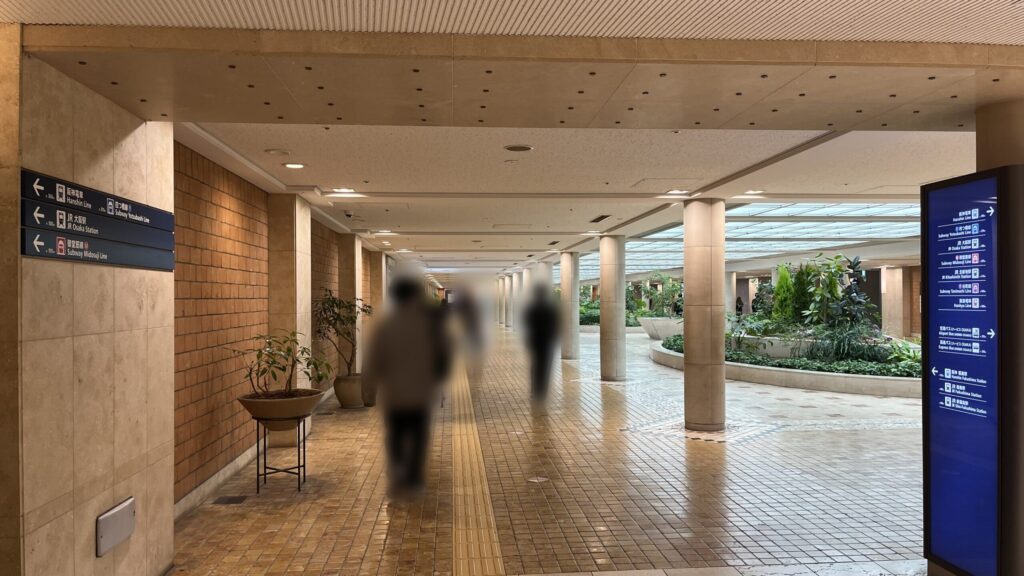SYNTH×Business-Airport 西梅田ブリーゼタワーへのアクセス方法：左右に路線案内板がある通路　白い円柱が並んでいるが、その右手に吹き抜けと緑の生い茂った噴水がある