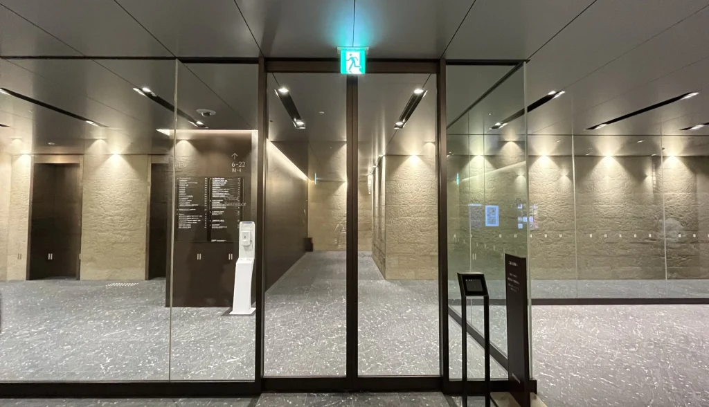 Signature大阪ダイビル本館へのアクセス方法：天井に緑色の非常口誘導灯が光る、全面ガラス張りのオフィスエントランスの自動扉が写っており、手前右側にはスタンド型の黒い端末が設置されています。