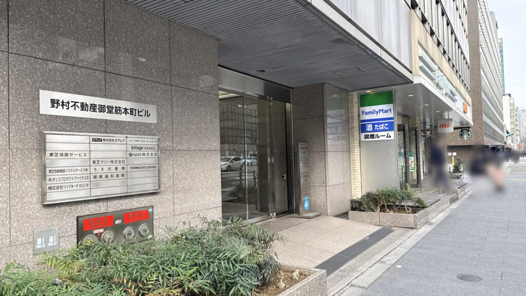 H¹O 本町Ⅱへの行き方：野村不動産御堂筋本町ビルのエントランス。ガラス張りの自動ドアがあり、左側の壁に「H¹O 本町Ⅱ」の黒い看板が設置されている。右隣にファミリーマートがある