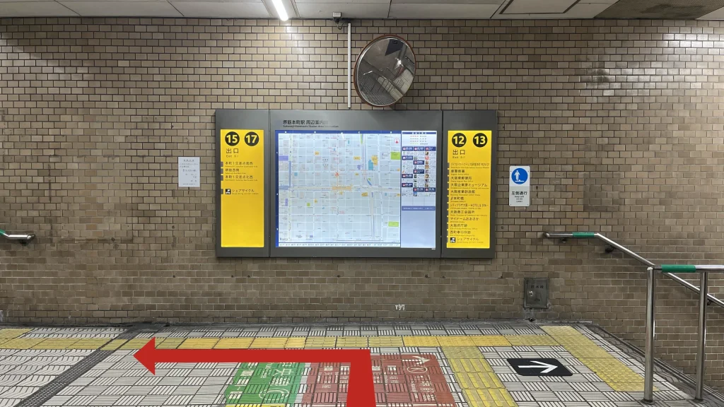 リージャス大阪国際ビルディングビジネスセンターへのアクセス方法：地下駅構内のタイル壁に設置された「15 17 出口」および「12 13 出口」と書かれた黄色の案内板と周辺地図。床面には白い矢印の入った案内タイルがある。