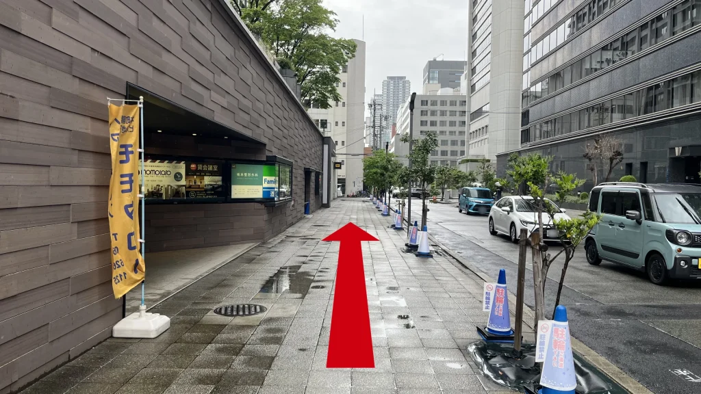 リージャス大阪国際ビルディングビジネスセンターへのアクセス方法：ビルの側面に沿った歩道。左側の壁面にはファミリーマートの看板やのぼりが設置されており、歩道には駐輪禁止 of コーンが並んでいる。