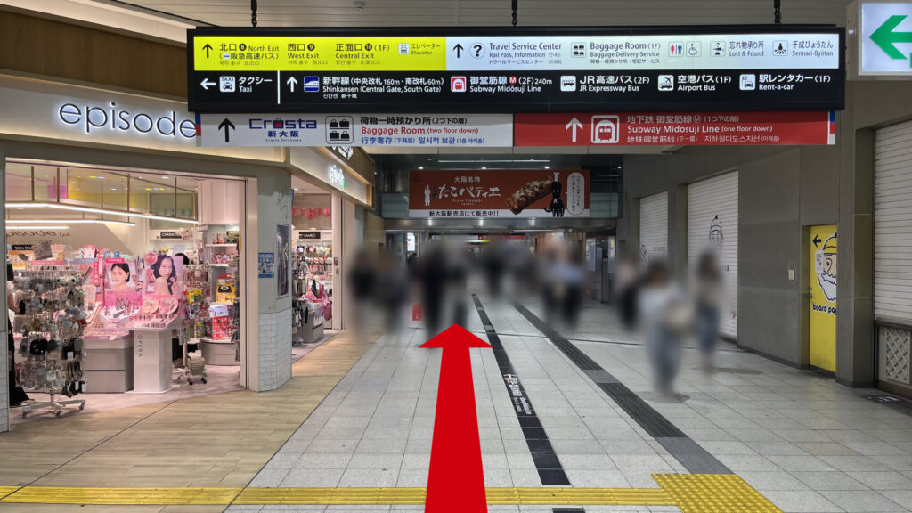 リージャス新大阪阪急ビルビジネスセンターへの行き方：店舗が並ぶ駅構内の広い通路。頭上には各方面への黄色と白の案内看板が設置されており、奥へと続く道を歩行者が進んでいる。