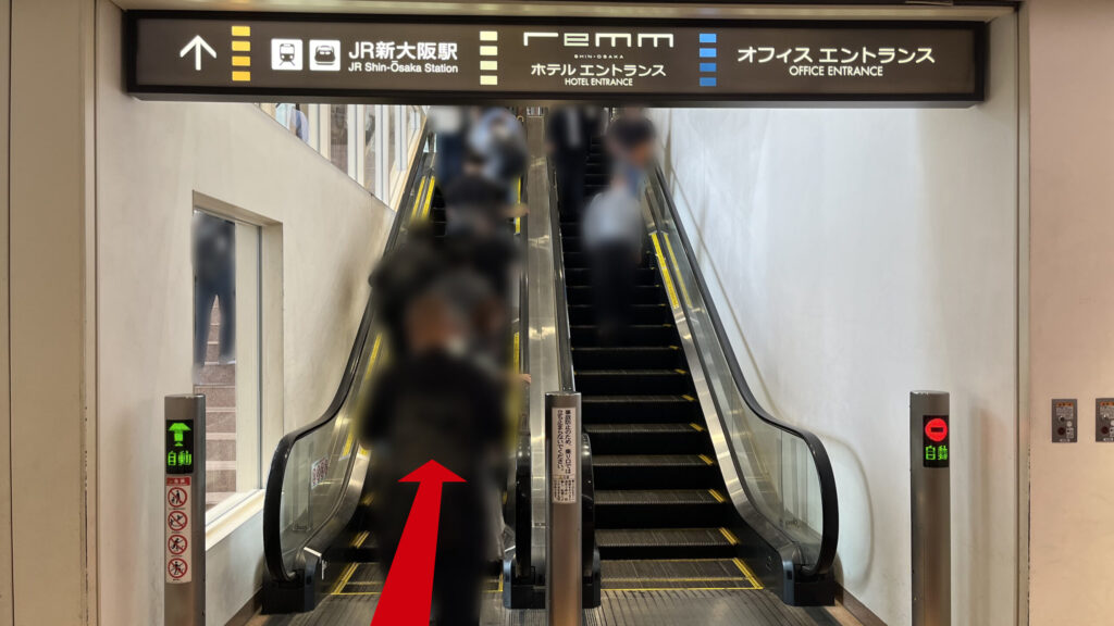 リージャス新大阪阪急ビルビジネスセンターへの行き方：ビル上層階へ続く上りエスカレーターの乗り口。頭上にはJR新大阪駅やオフィスエントランスへと導く黒い案内板が掲げられている。