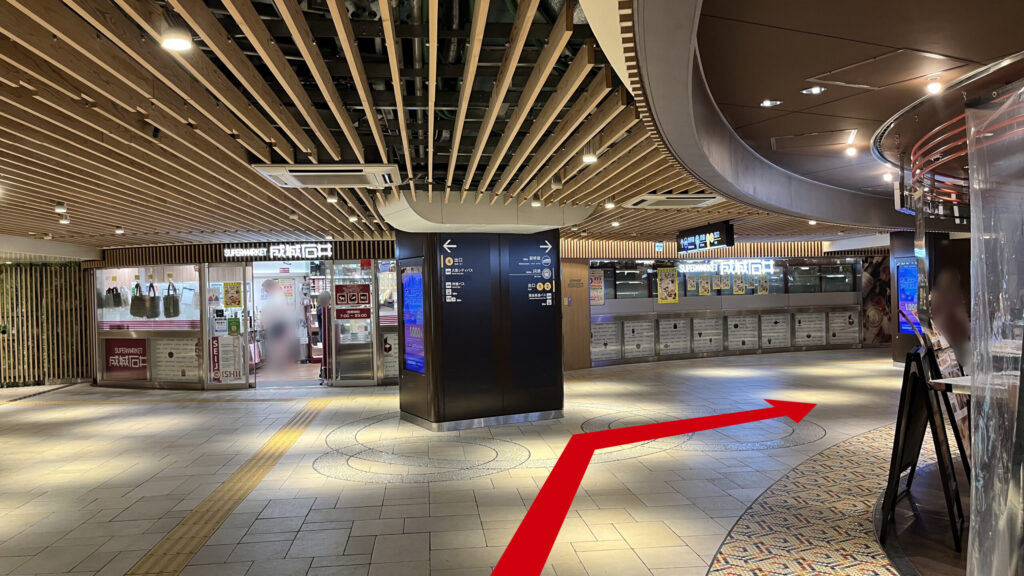リージャス新大阪阪急ビルビジネスセンターへの行き方：木目のルーバー天井が続く地下通路。左奥には成城石井の店舗があり、中央に置かれた案内柱の右側を抜けるように通路が延びている。