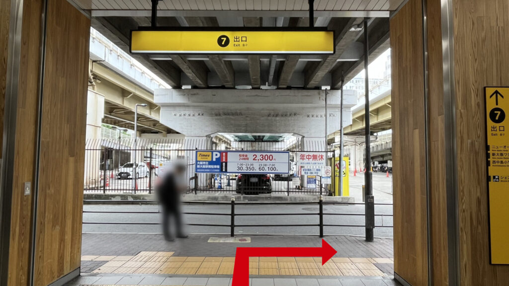 リージャス新大阪南ビジネスセンターへの行き方：７番出口付近の写真　頭上と右側に７番出口の黄色い看板、歩道の反対側に駐車場が広がっており、上には新御堂筋が走っている